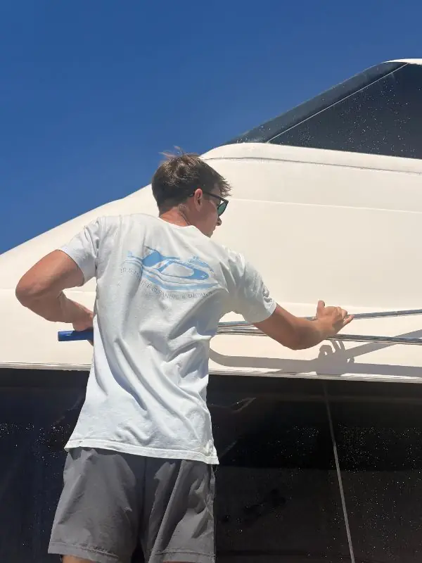Social post photo: a young man in a JD's Marine Cleaning & Detailing t-shirt cleaning the hull of a white boat under a blue sky
